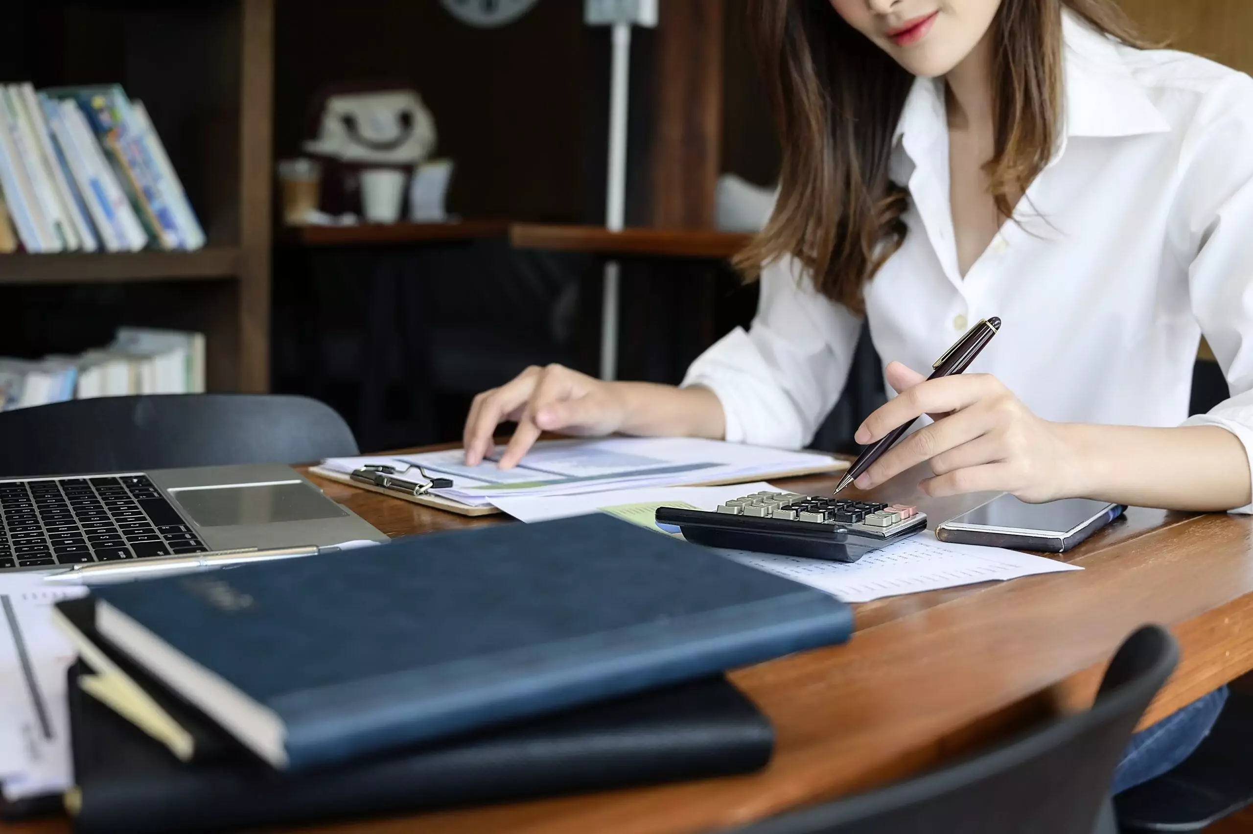 Cropped shot of young woman holding pen and using calculator while sitting at office desk.